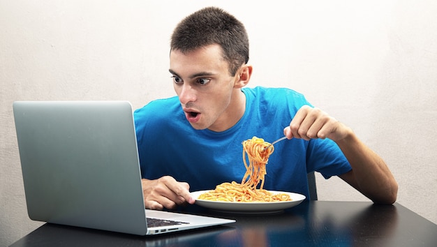 Jeune homme mangeant des spaghettis à la sauce tomate et regardant l'ordinateur