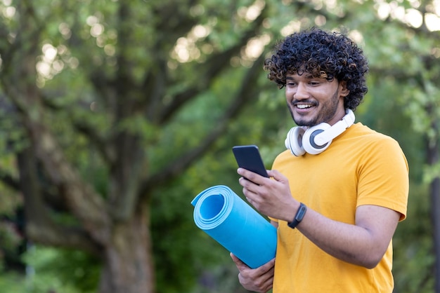 Jeune homme marchant dans le parc pour l'entraînement de yoga homme hispanique avec tapis de fitness agrandi avec un casque