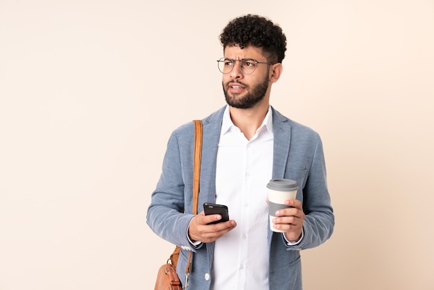 Jeune homme marocain d'affaires isolé sur mur beige tenant du café à emporter et un mobile tout en pensant à quelque chose