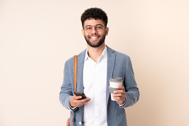 Jeune homme marocain d'affaires isolé sur mur beige tenant du café à emporter et un mobile