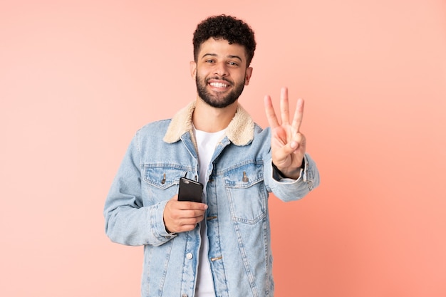 Jeune homme marocain à l'aide de téléphone mobile isolé sur fond rose heureux et en comptant trois avec les doigts