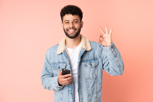 Jeune homme marocain à l'aide de téléphone mobile isolé sur fond rose montrant signe ok avec les doigts