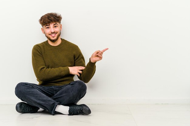 Jeune homme marocain assis sur le sol isolé sur blanc souriant joyeusement pointant avec l'index loin.