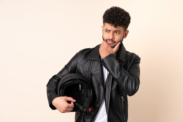 Jeune homme marocain avec un casque de moto isolé sur fond beige ayant des doutes