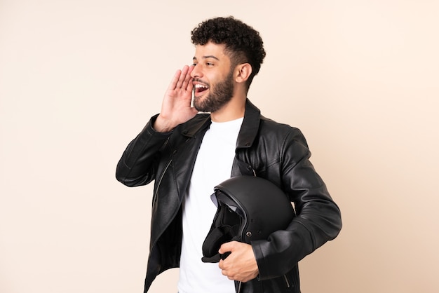 Jeune homme marocain avec un casque de moto isolé sur un mur beige en criant avec la bouche grande ouverte sur le côté