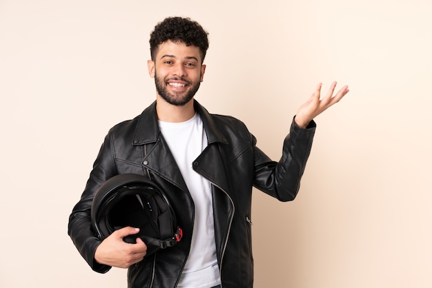 Jeune homme marocain avec un casque de moto isolé sur mur beige étendant les mains sur le côté pour inviter à venir