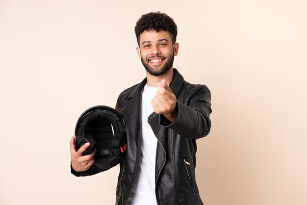 Jeune homme marocain avec un casque de moto isolé sur un mur beige faisant un geste d'argent