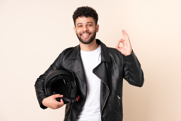 Jeune homme marocain avec un casque de moto isolé sur mur beige montrant signe ok avec les doigts