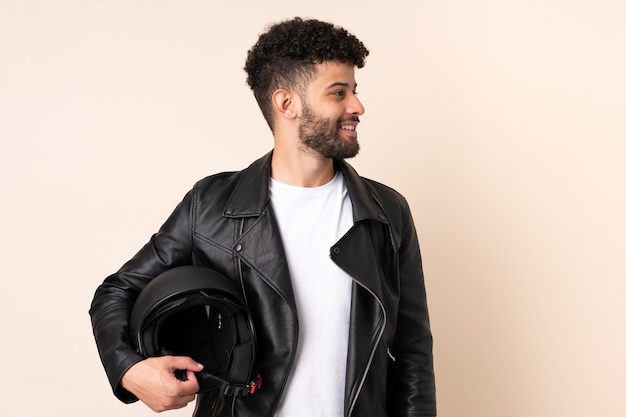 Jeune homme marocain avec un casque de moto isolé sur un mur beige à la recherche sur le côté