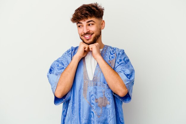 Jeune homme marocain isolé sur blanc garde les mains sous le menton, regarde joyeusement de côté.