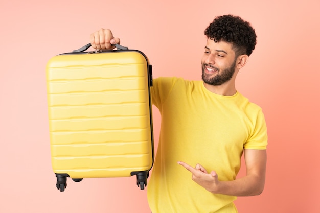 Jeune homme marocain isolé sur un mur rose en vacances avec valise de voyage