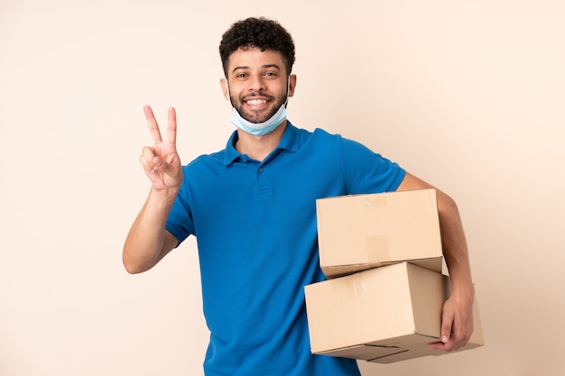 Jeune homme marocain de livraison isolé sur beige souriant et montrant le signe de la victoire