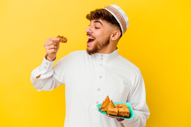 Jeune homme marocain portant le costume arabe typique de manger des bonbons arabes isolé sur mur jaune