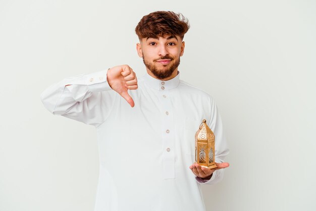 Jeune homme marocain portant des vêtements arabes typiques tenant une lampe de ramadan isolé sur blanc montrant un geste d'aversion, les pouces vers le bas