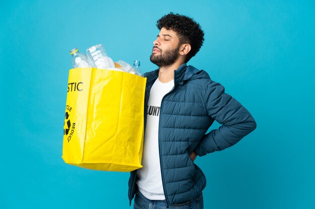 Jeune homme marocain tenant un sac plein de bouteilles en plastique à recycler sur fond isolé souffrant de maux de dos pour avoir fait un effort