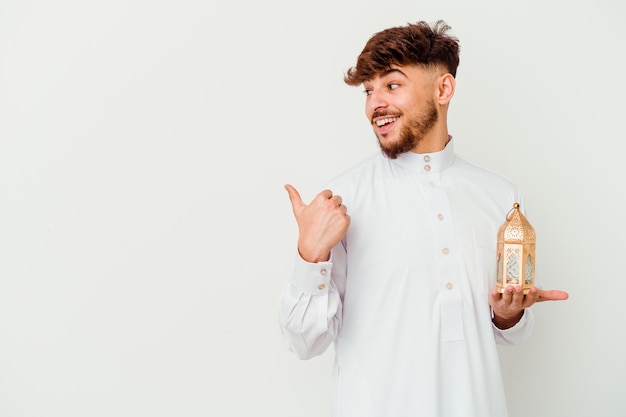 Jeune homme marocain vêtu d'un vêtement arabe typique tenant une lampe de ramadan isolée sur des points blancs avec le pouce, riant et insouciant.