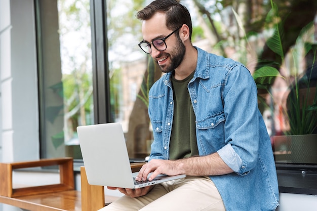 jeune homme masculin portant des lunettes en tapant sur un ordinateur portable tout en travaillant dans un café de la ville à l'extérieur