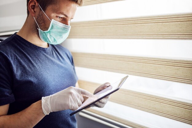 Jeune homme avec masque et gants tapant sur tablette, à la recherche de nouvelles en ligne. Soyez responsable, restez à la maison. Pandémie mondiale.