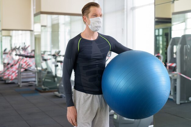 Jeune homme avec masque pensant tout en tenant un ballon d'exercice au gymnase pendant le coronavirus covid-19