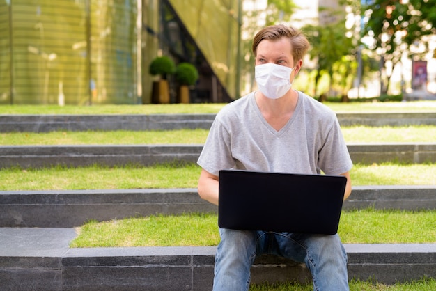Jeune homme avec masque pensant et utilisant un ordinateur portable alors qu'il était assis à l'extérieur