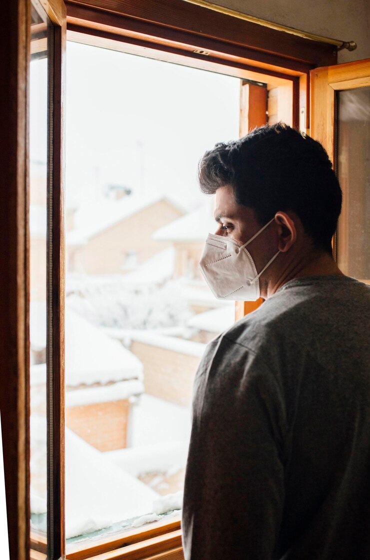 Photo un jeune homme avec un masque en quarantaine regardant la chute de neige à travers la fenêtre de sa maison