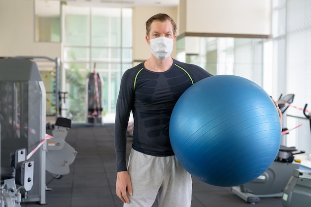 Jeune homme avec masque tenant un ballon d'exercice au gymnase pendant le coronavirus covid-19