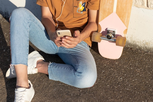 Jeune homme méconnaissable assis sur le sol de la ville écoutant de la musique et envoyant des SMS et une planche à roulettes sur le côté