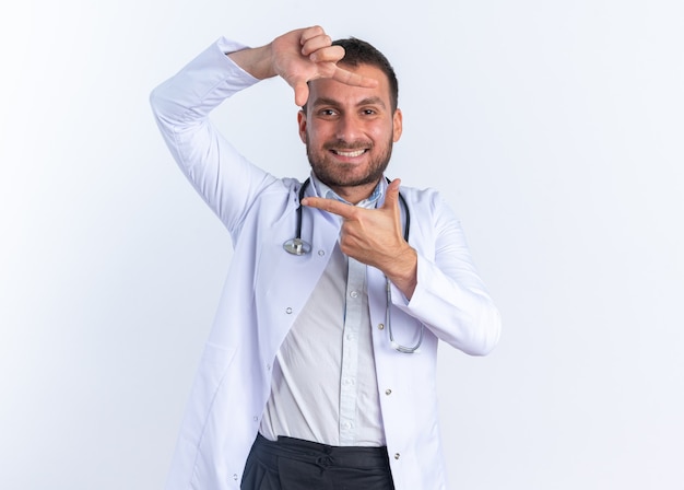 Jeune homme médecin en blouse blanche et avec stéthoscope autour du cou faisant cadre avec les doigts regardant à travers ce cadre souriant joyeusement