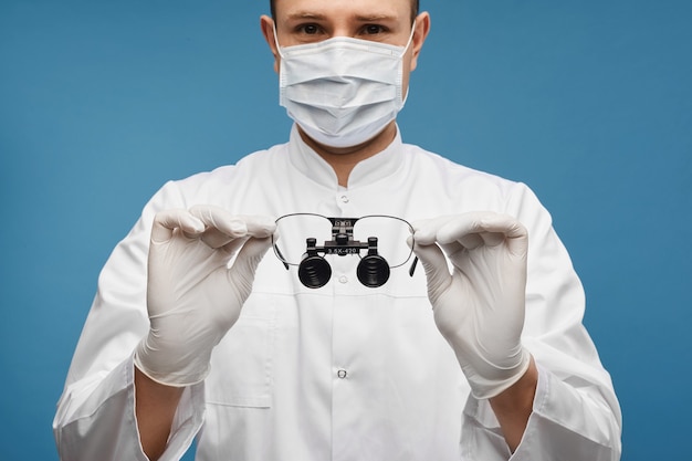 Jeune homme médecin dans un masque chirurgical et des gants de protection garde des loupes binoculaires dans ses mains, isolé sur le fond bleu avec copie espace