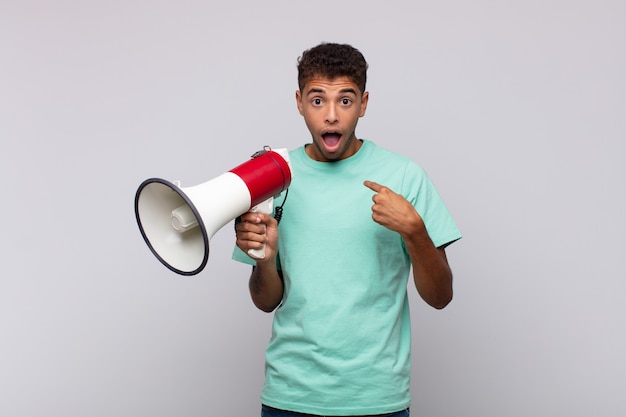 Jeune homme avec un mégaphone se sentant heureux, surpris et fier, pointant vers soi avec un regard excité et étonné