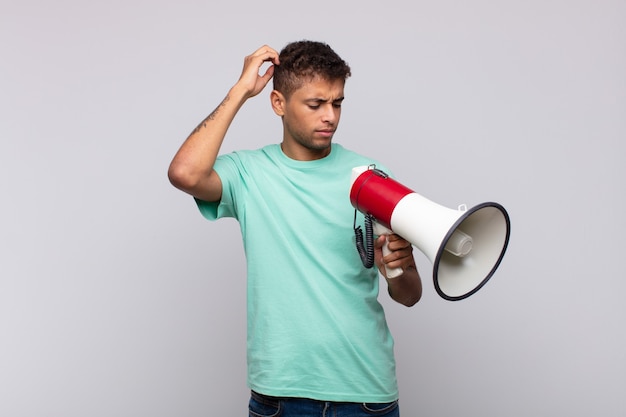 Jeune homme avec un mégaphone se sentant perplexe et confus, se gratte la tête et regarde sur le côté