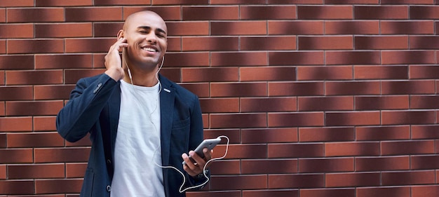 Photo jeune homme mélomane portant des écouteurs debout sur le mur écoutant de la musique sur un smartphone fermé