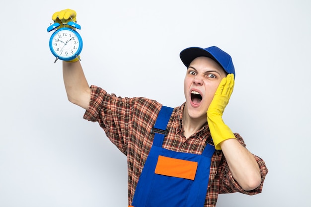 Jeune Homme De Ménage Portant Un Uniforme Et Une Casquette Avec Des Gants Tenant Un Réveil Isolé Sur Un Mur Blanc