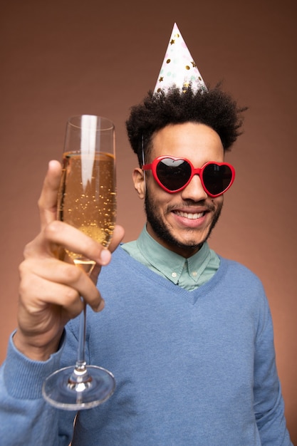 Jeune homme métis gai dans une casquette d'anniversaire et des lunettes de soleil en forme de coeur grillant avec une flûte de champagne devant la caméra