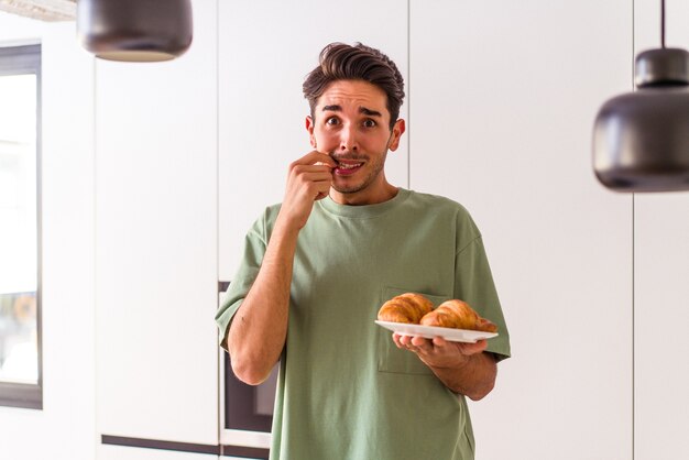 Jeune homme métis mangeant un croissant dans une cuisine le matin se rongeant les ongles, nerveux et très anxieux.