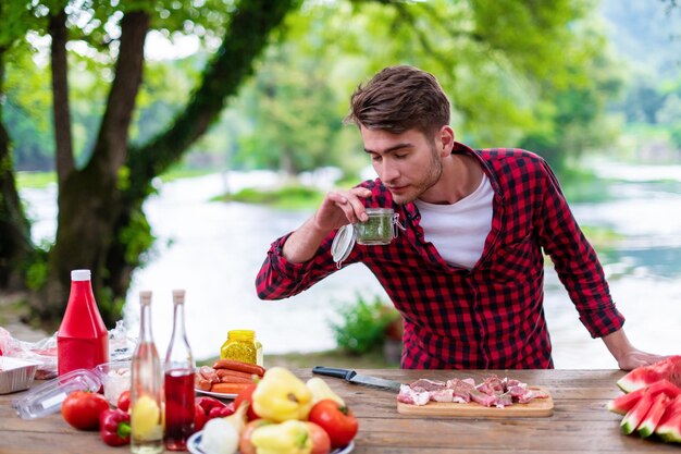 jeune homme mettant des épices sur de la viande crue pour un barbecue lors d'un dîner français en plein air près de la rivière lors d'une belle soirée d'été dans la nature