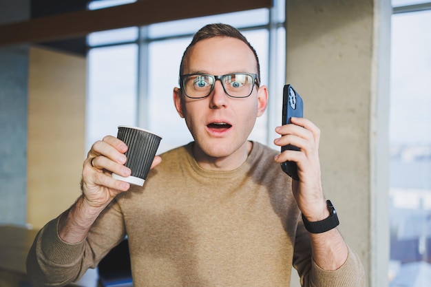 Un jeune homme mignon dans un pull marron et des lunettes parle avec émotion sur un téléphone portable et boit du café tout en se relaxant au bureau Un jeune pigiste travaille à distance