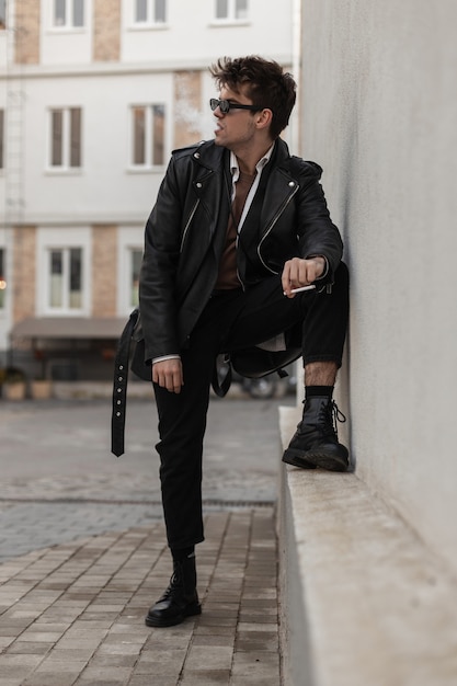 Jeune homme à la mode hipster dans des vêtements noirs surdimensionnés élégants dans des lunettes de soleil reposant avec une cigarette près d'un bâtiment vintage dans la rue. Un beau mec brutal dans des vêtements de jeunesse branchés fume à l'extérieur.