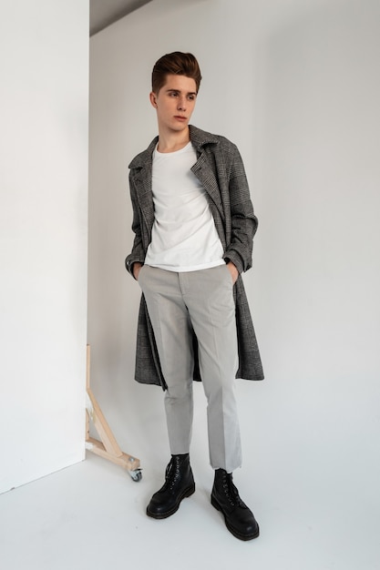 Jeune homme à la mode en T-shirt blanc en manteau à carreaux tendance dans un pantalon élégant en bottes noires en cuir à la mode se tient près du mur en studio. Beau modèle de mec moderne en vêtements d'extérieur décontractés à la mode dans la chambre