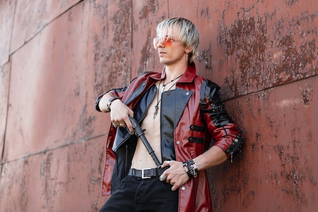 Jeune homme à la mode en veste en cuir noir-rouge élégante dans des lunettes à la mode torse nu dans un jean avec un pistolet vintage pose près du mur rouillé en métal à l'extérieur.