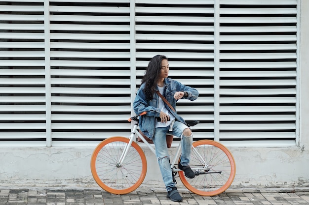 Jeune homme à la mode en vêtements en jean s'appuyant sur un vélo et vérifiant l'heure ou les notifications sur son ...