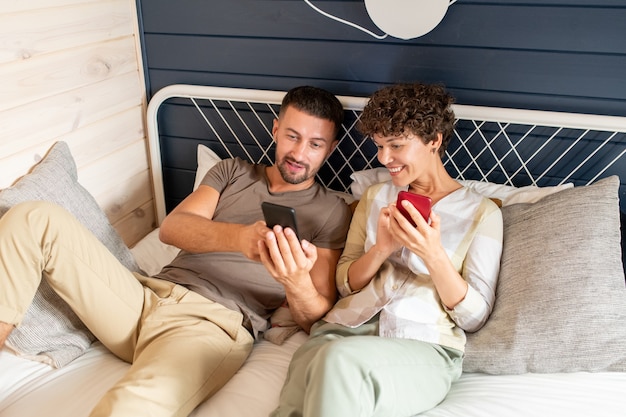 Jeune homme montrant à sa femme heureuse une vidéo en ligne curieuse ou une publication sur les réseaux sociaux sur un smartphone tout en se relaxant sur le lit à loisir