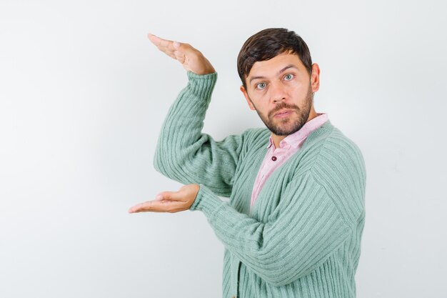 Jeune homme montrant un signe de grande taille en chemise, cardigan et semblant concentré, vue de face.