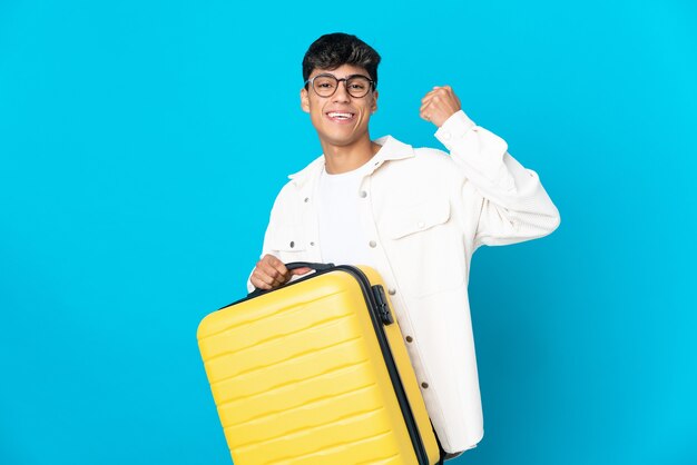 Jeune homme sur mur bleu isolé en vacances avec valise de voyage