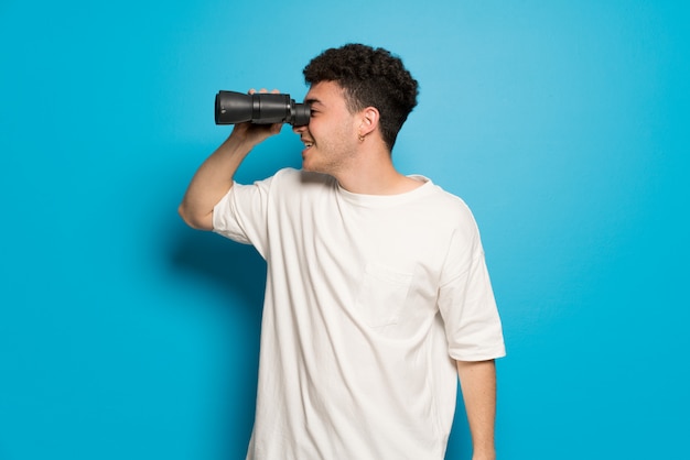 Jeune homme sur un mur bleu et regardant au loin avec des jumelles