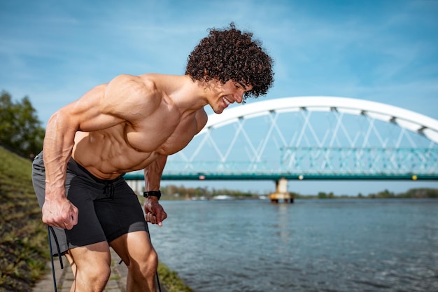 Un jeune homme musclé au torse nu fait un entraînement corporel solide avec un élastique, au bord de la rivière.