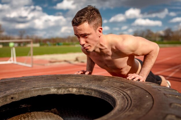 Jeune homme musclé faisant des pompes sur un énorme pneu pour entraîner les muscles sur la piste en tartan du stade.