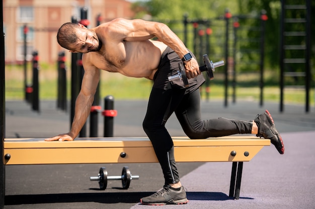 Jeune homme musclé en leggins noirs s'appuyant sur un banc tout en soulevant des haltères lourds sur le terrain de sport