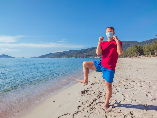 Un jeune homme musclé portant un masque médical effectue des entraînements en plein air dans le parc près de la mer pendant la deuxième vague de coronavirus Covid 19 quarantaine pandémique Kick training pose bras et jambes oscillants sport vie active