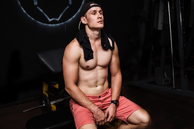 Jeune homme musclé et torse nu au repos dans la salle de sport pendant l'entraînement, montrant le torse musclé, les pectoraux et les abdominaux, assis sur un banc